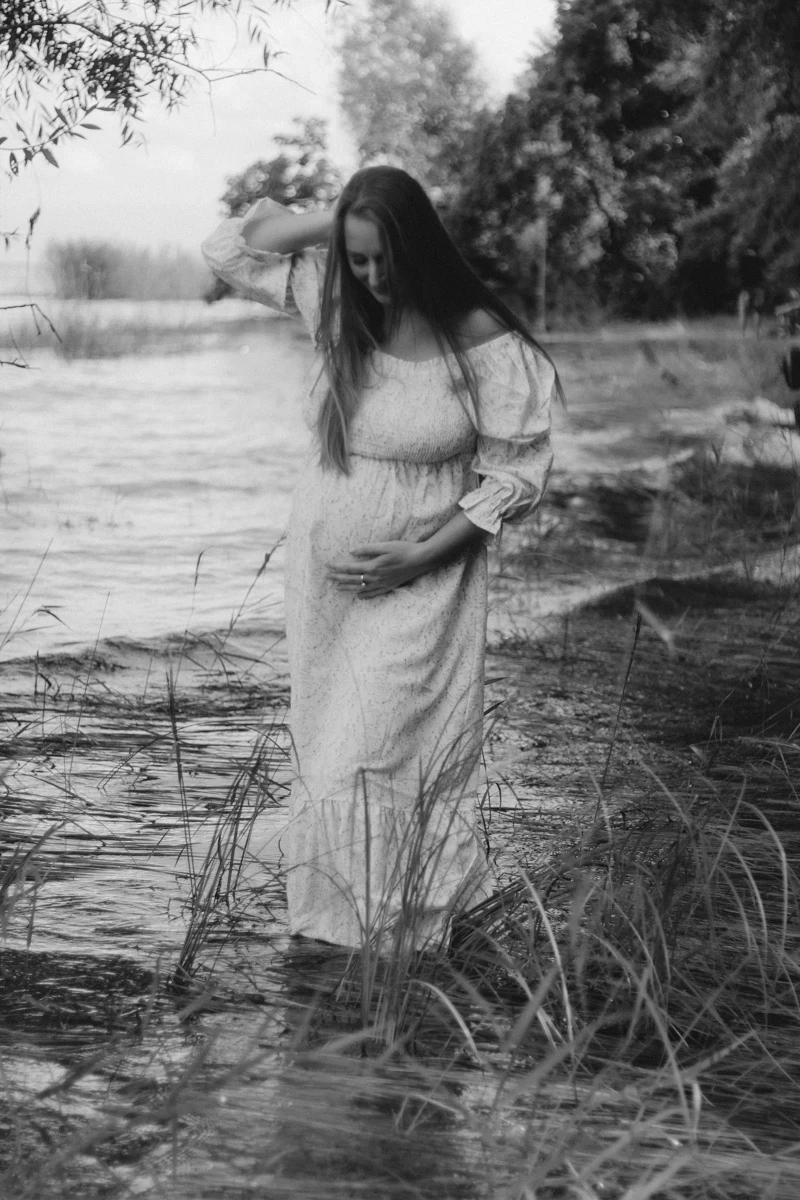 Eine schwangere Frau steht in romantischem Kleid mit den Füßen im Wasser und betrachtet ihren Bauch