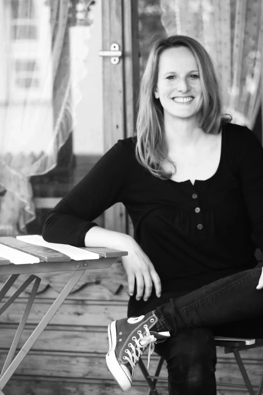 Hier sehen Sie die Fotoartistin Barbara Kremer mit mittellangen, glatten Haaren. Sie trägt eine schwarze Bluse und schwarze Jeans. Sie sitzt an einem kleinen, gestreiften Tisch im Freien. An ihren Füßen trägt sie schwarz-weiße Converse Turnschuhe mit weißen Schnürsenkeln. Der Hintergrund deutet auf eine entspannte, häusliche Umgebung hin, möglicherweise eine Terrasse oder ein Balkon, mit hellen Vorhängen im Unschärfebereich.