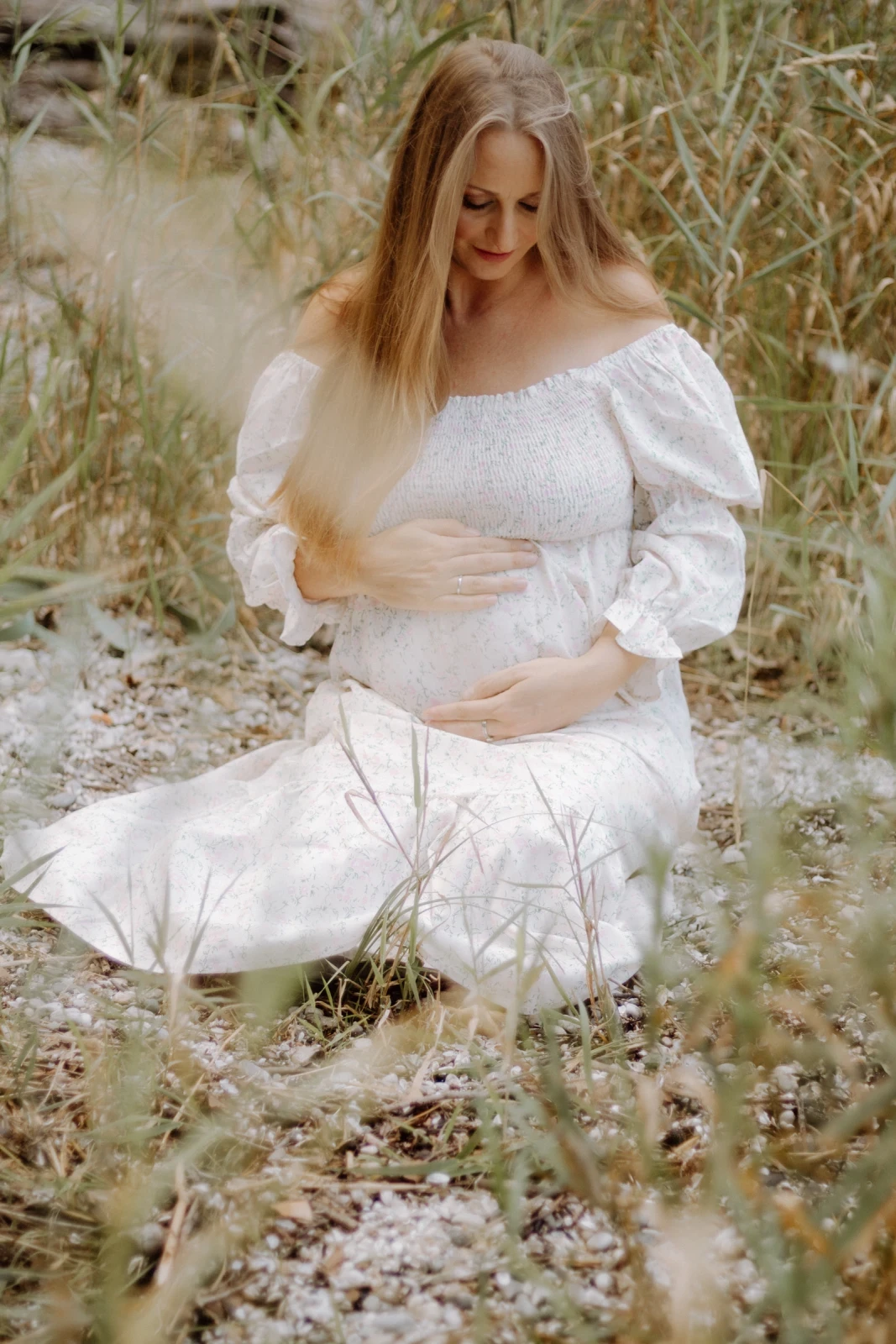 Eine schwangere Frau mit einem romantischem Boho - Kleid sitzt gedankenversunken auf dem Boden und betrachtet ihren Bauch