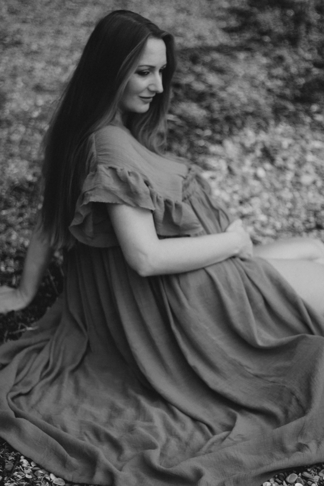 Schwangere Frau sitzt im romantischen Boho-Kleid am Ufer und berührt nachdenklich ihren Bauch