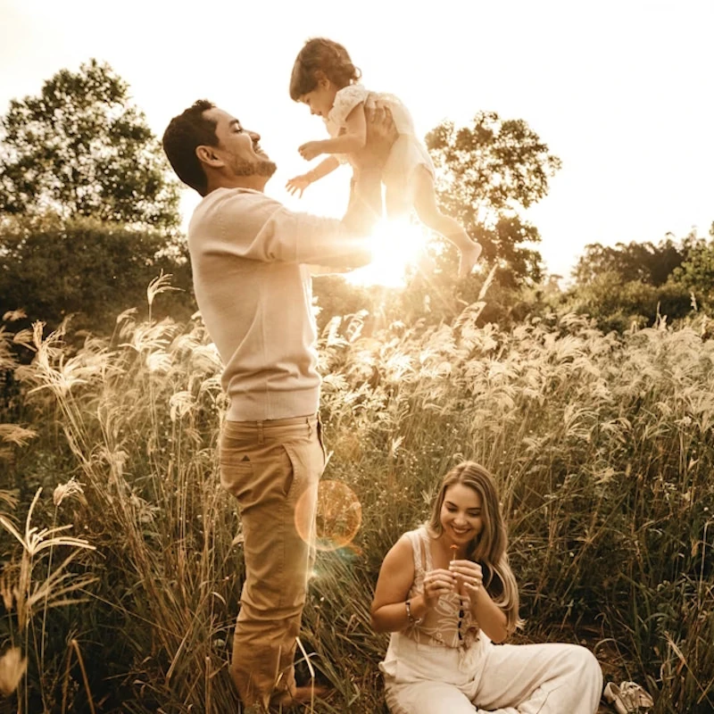 Eine glückliche Familie von fünf Personen – Vater, Mutter und drei Kinder – spaziert Hand in Hand durch ein hohes, grünes Grasfeld. Die Mutter trägt ein langes, rotes Kleid mit weißen Blüten und einen Strohhut, während sie mit dem älteren Jungen und dem Vater, der ein Baby auf dem Arm hält, verbunden ist. Der jüngere Junge in Hosenträgern geht zwischen seinen Geschwistern. Im Hintergrund, hinter einem niedrigen Zaun, sind Bäume und eine Lichterkette in der Dämmerung zu sehen.